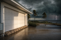 Gulf coastal home with burlap StormBag flood bags along the garage door, wind-bent palms and an approaching tropical storm with lightning over the Gulf of Mexico